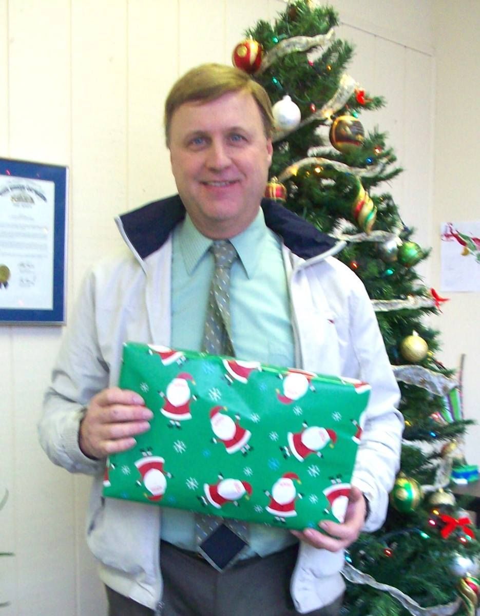 Man holding a Christmas present wrapped in green paper with Santa Claus print; Christmas tree in the background.