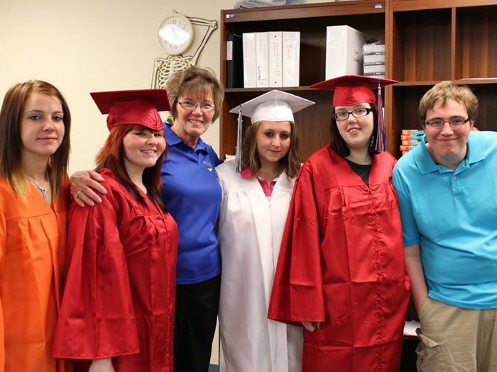 Group of graduates in red and white gowns with a woman in a blue shirt posing for a photo in a room with bookshelves.