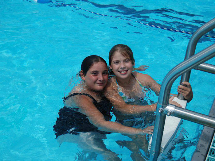 Two girls in a swimming pool, smiling. One holds pool ladder. Bright blue water.