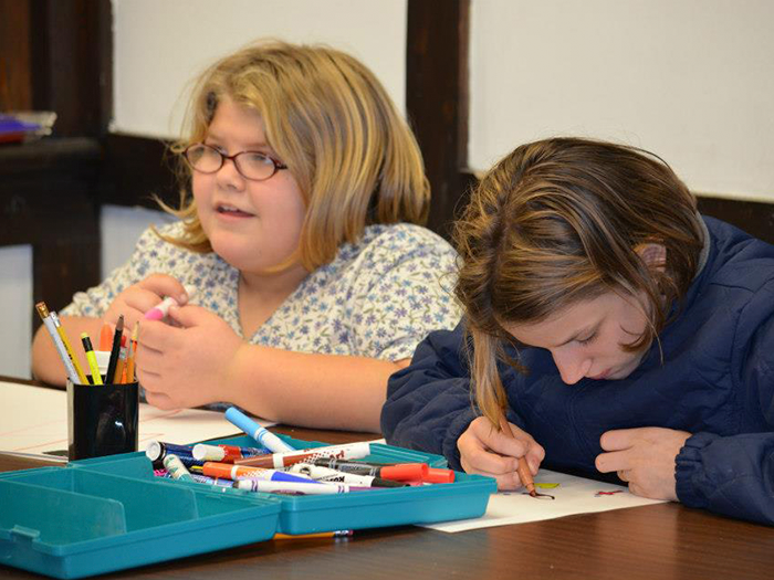Two people drawing with markers at a table. One wears glasses and looks to the side, the other is focused on their work.