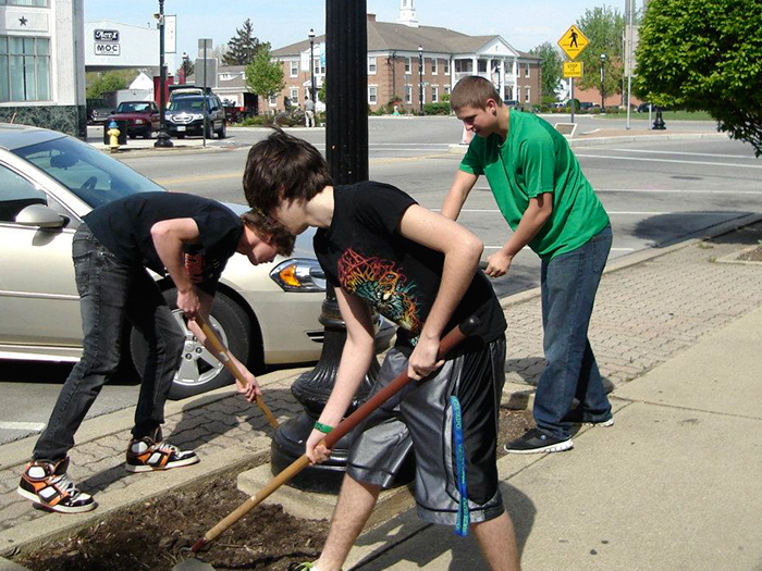 Three people working outdoors, using tools to tend to a flower bed by a sidewalk near a street.