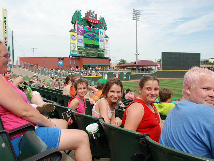People watching baseball game in stadium; scoreboard in background.