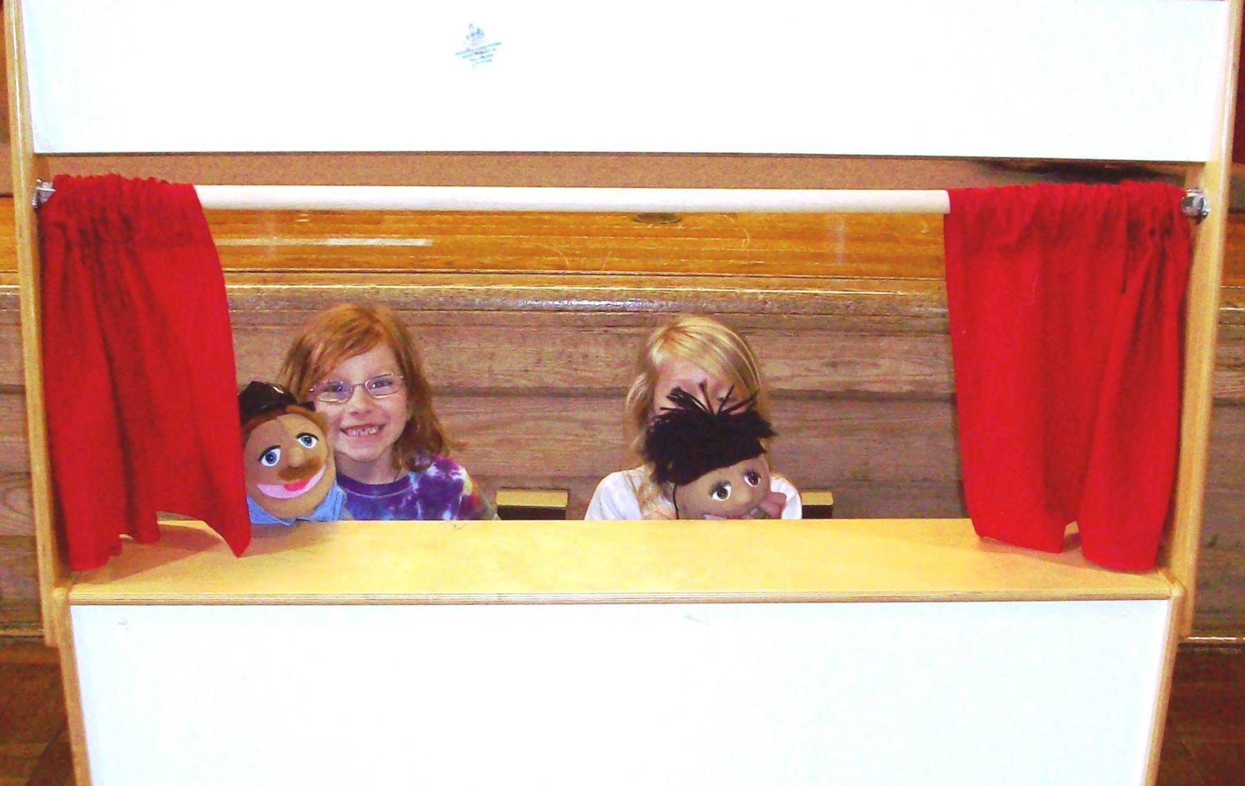 Children performing a puppet show, using a small stage with red curtains.