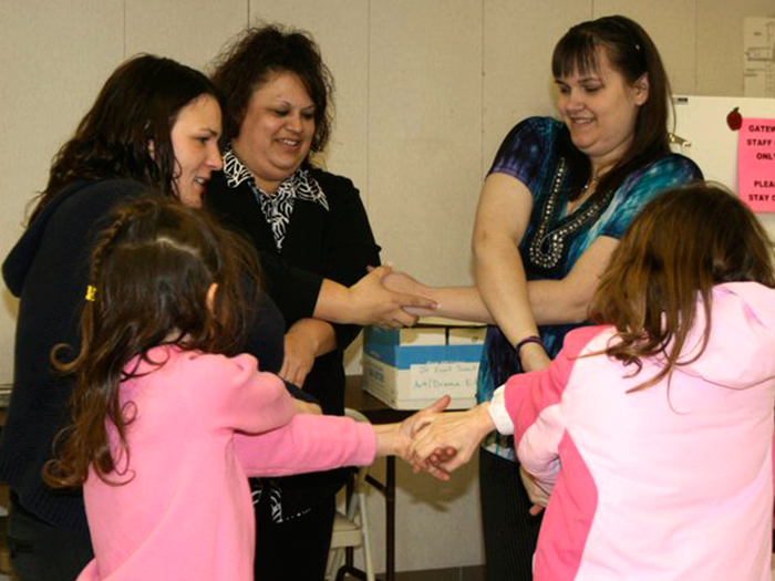 Five people holding hands in a circle, smiling, indoors. Two children wear pink, adults dark and light tops.