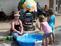 People at an outdoor event; a woman with a balloon hat sits by a small pool, kids play nearby, balloons overhead.