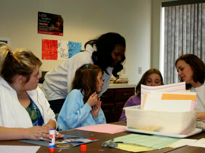 Group of people gathered around a table; crafting with paper. One person is giving instructions.