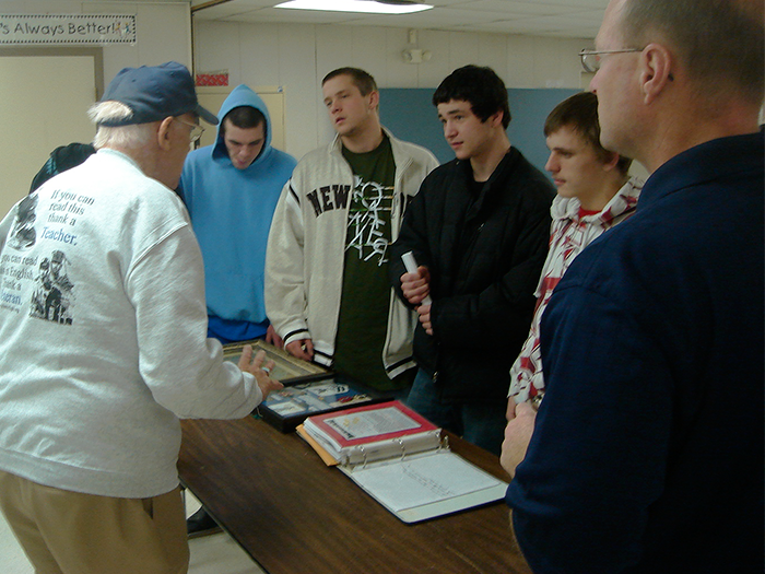 A man shows documents to a group of students. They stand around a table in a classroom.