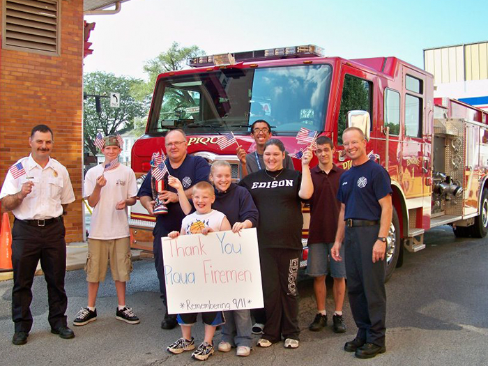 People with firefighters and fire truck, holding American flags and sign.