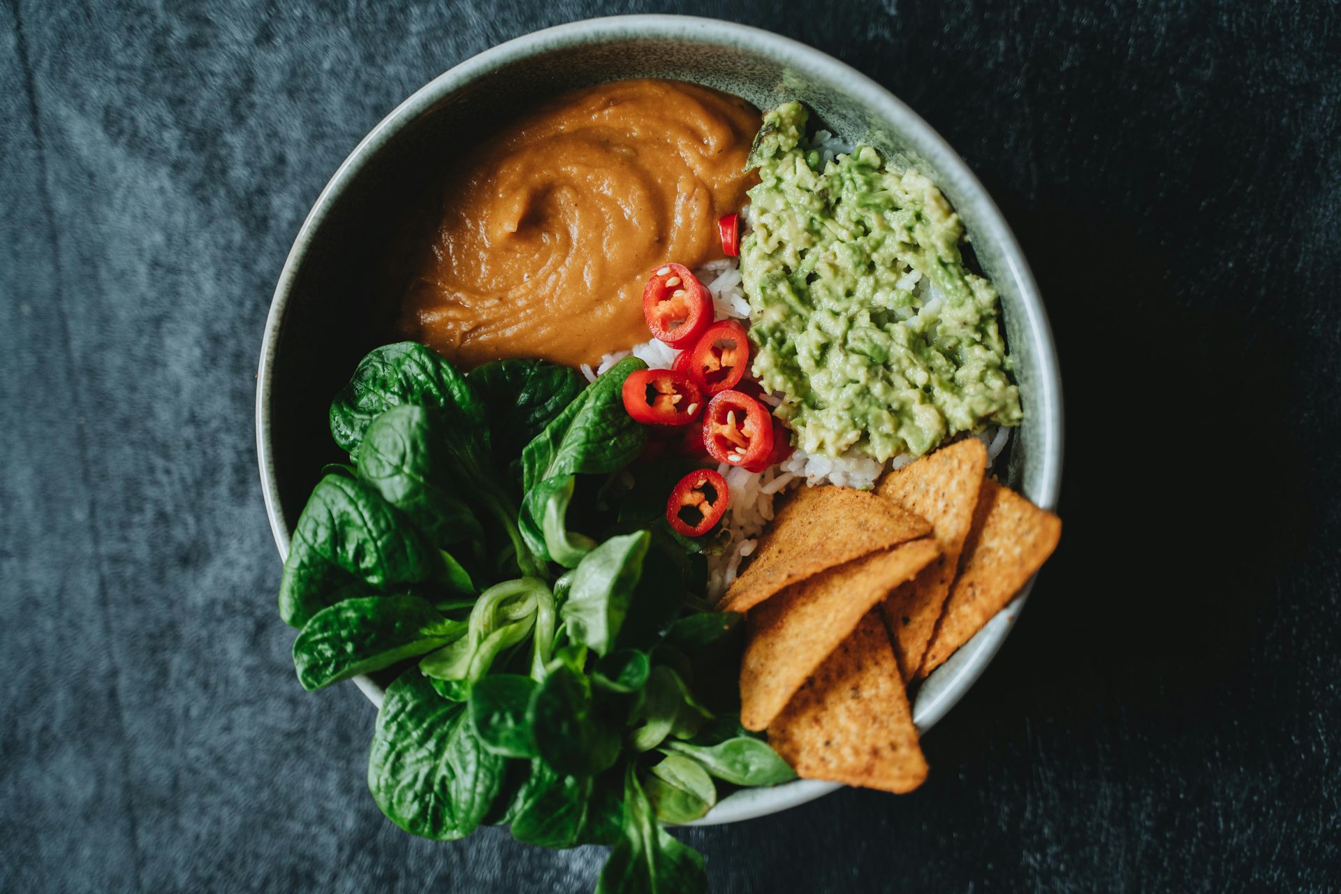 Cuenco con espinacas, salsa de maní, aguacate, arroz, pimientos y patatas fritas sobre una superficie oscura.