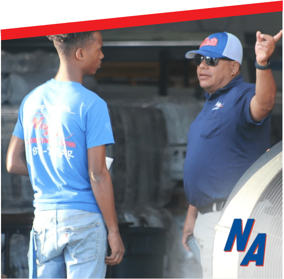 Two men talking in a garage, one wearing a blue shirt and cap with his hand raised.
