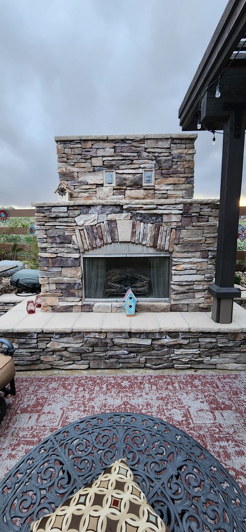 Stone outdoor fireplace on a patio with a patterned rug and furniture under an overcast sky.