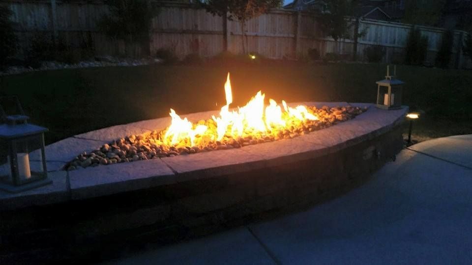 Fire pit with flames at dusk, surrounded by stone and lights in a backyard setting.