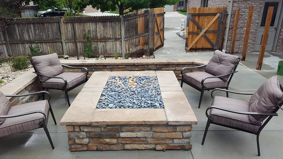 A stone fire pit with blue glass rocks, surrounded by four chairs on a patio with a wooden fence.