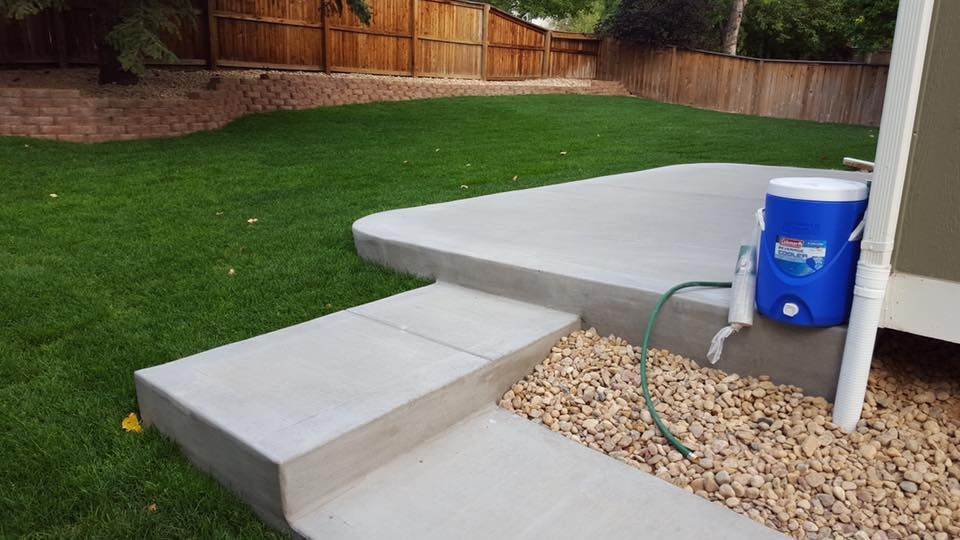 Concrete patio with steps, lush green grass, and a wooden fence. A blue water cooler sits on the patio.