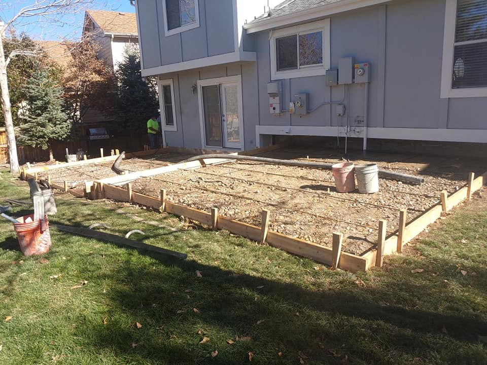 Construction site. Wooden forms outline a gravel-filled area near a house. Rebar and a bucket are present.