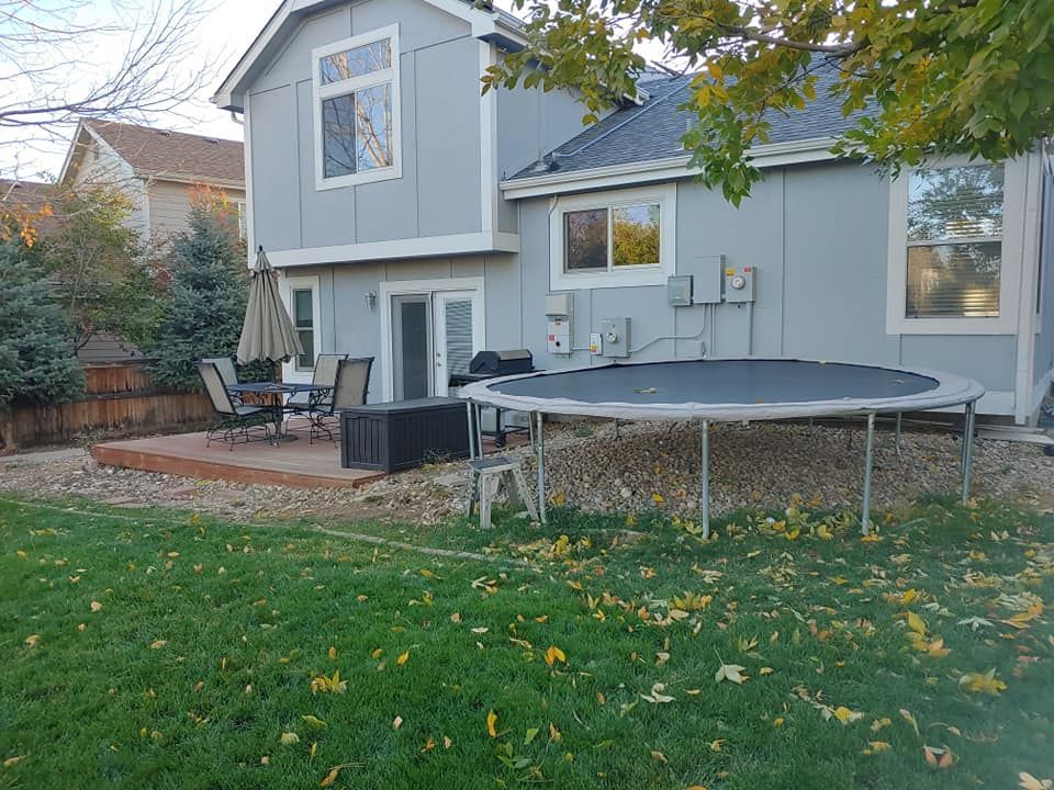 Backyard with a light blue house, trampoline, patio furniture, and green grass.