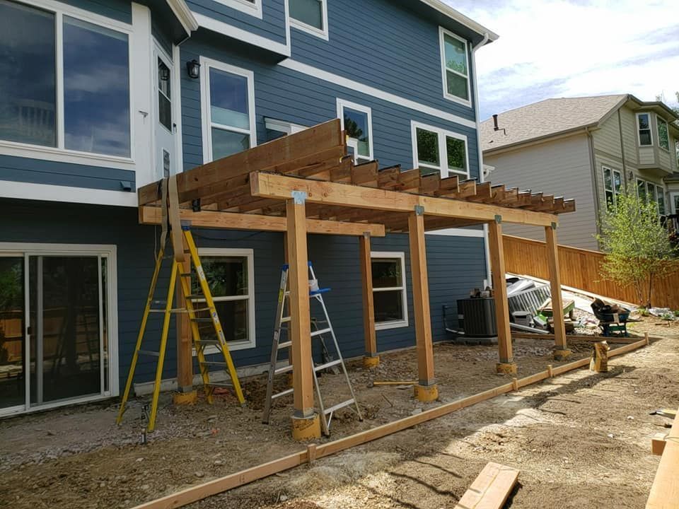 A partially constructed wooden pergola attached to a blue house, with ladders and construction materials.