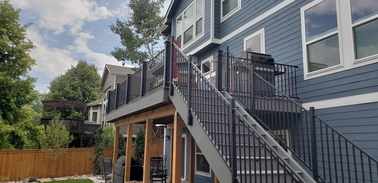 A two-story blue house with an attached deck and stairs. A wooden fence and trees in the background.