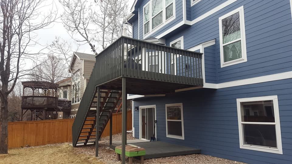A two-story blue house with a wooden deck and staircase. A wooden fence and yard are visible.