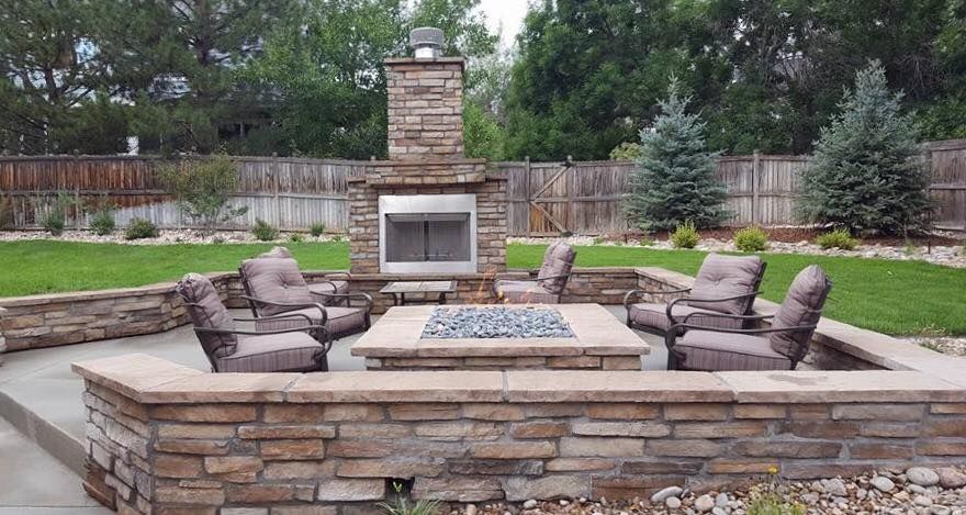 Outdoor seating area with stone fireplace, fire pit, and armchairs. Green lawn, trees, and wooden fence in the background.