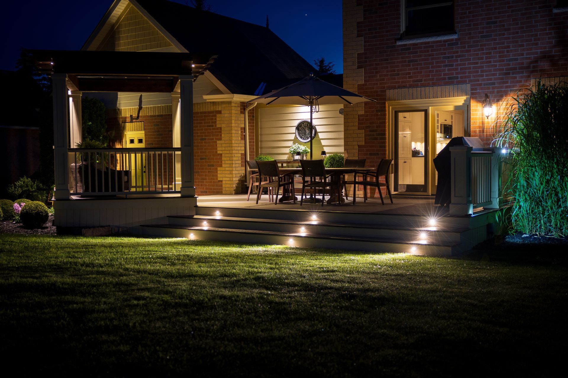 Night view of a backyard patio with lights illuminating steps, furniture, and a house.