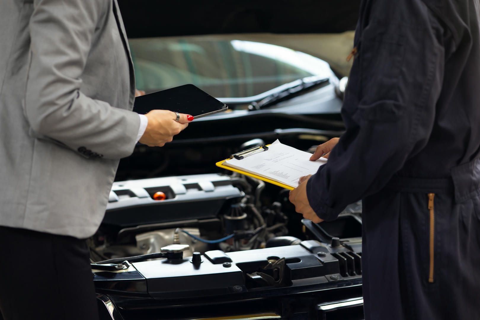 Car mechanic and client reviewing paperwork at an open car hood, outdoors.