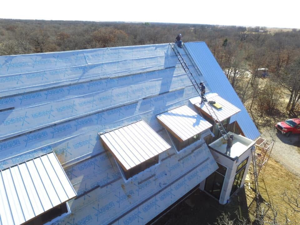 Worker on a ladder repairing a blue metal roof with equipment and materials nearby
