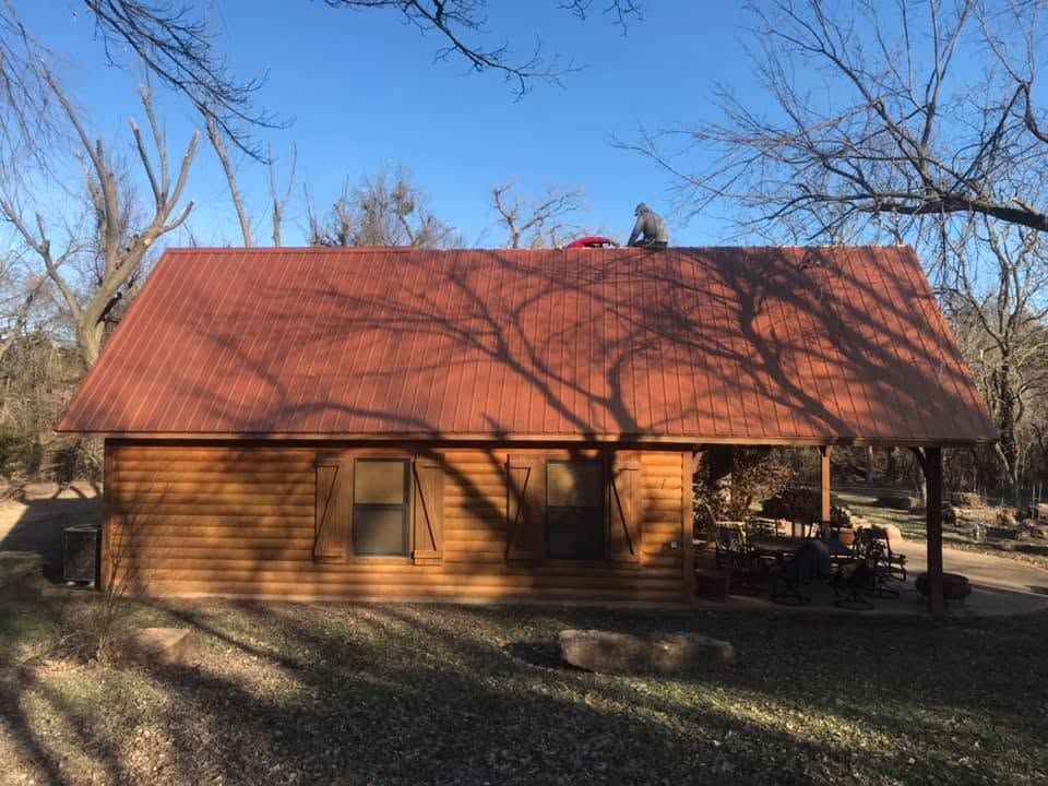 Small wooden cabin with a red roof and porch, surrounded by bare trees in a rural yard.