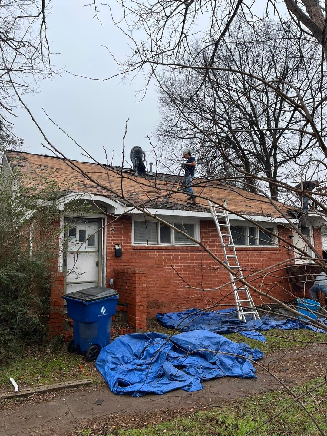 Brick house with workers on roof, ladder leaning up, and blue tarps covering the yard