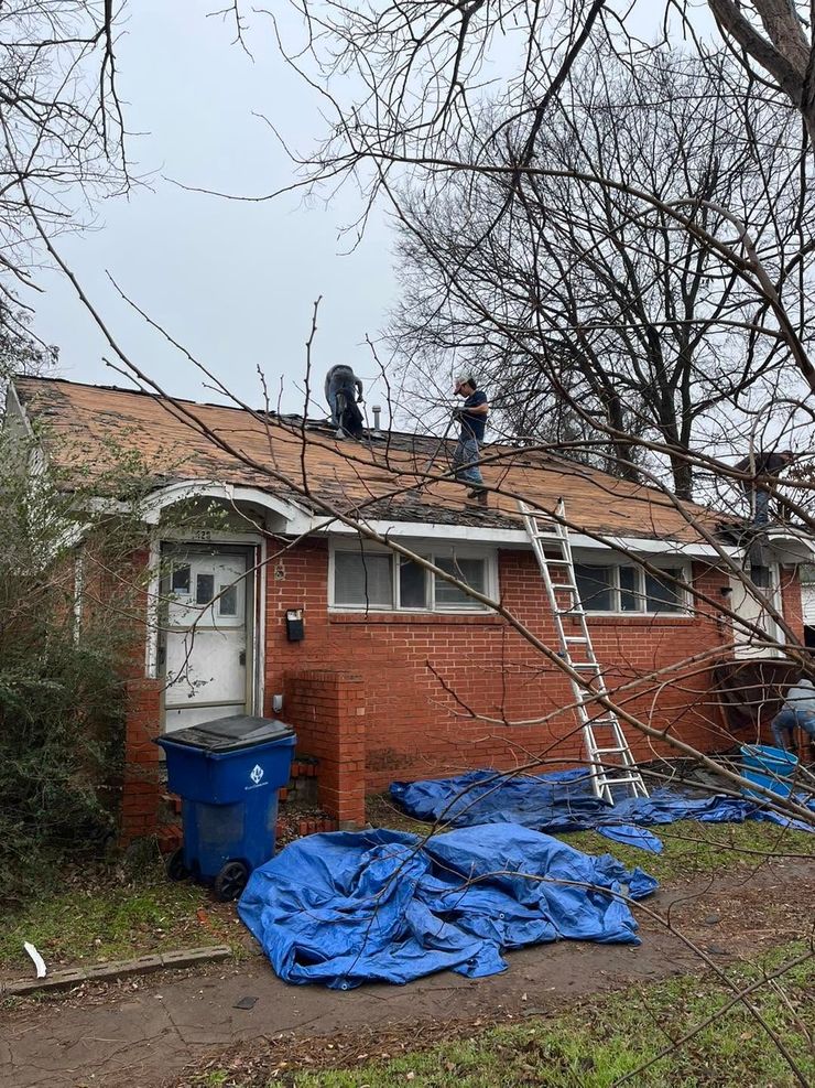 Workers on a brick house roof with ladders, blue tarps, and debris in the yard
