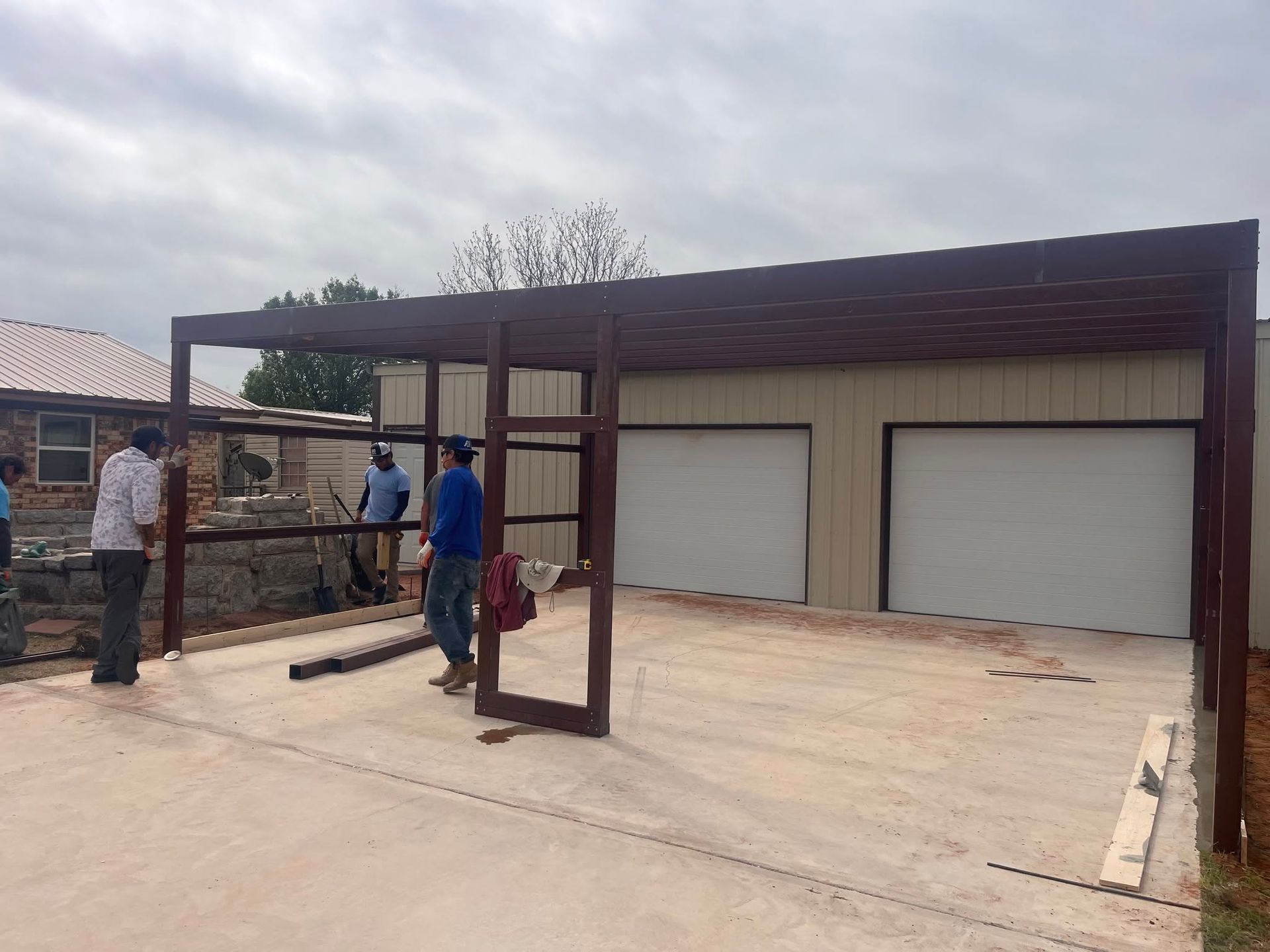 Workers installing a metal carport frame beside a garage with two white doors