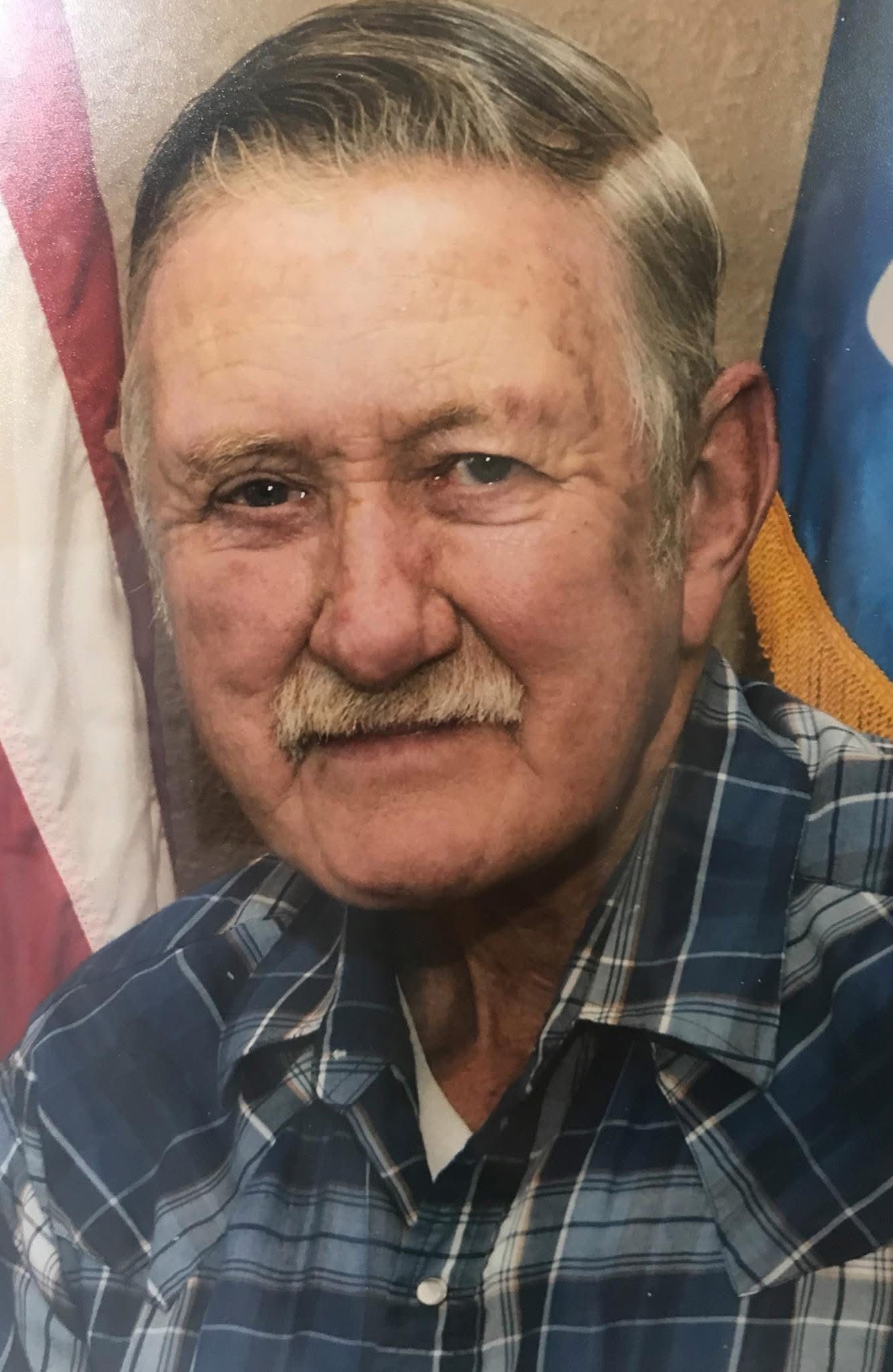 Portrait of a man in a blue plaid shirt, standing before U.S. and state flags with a slight smile