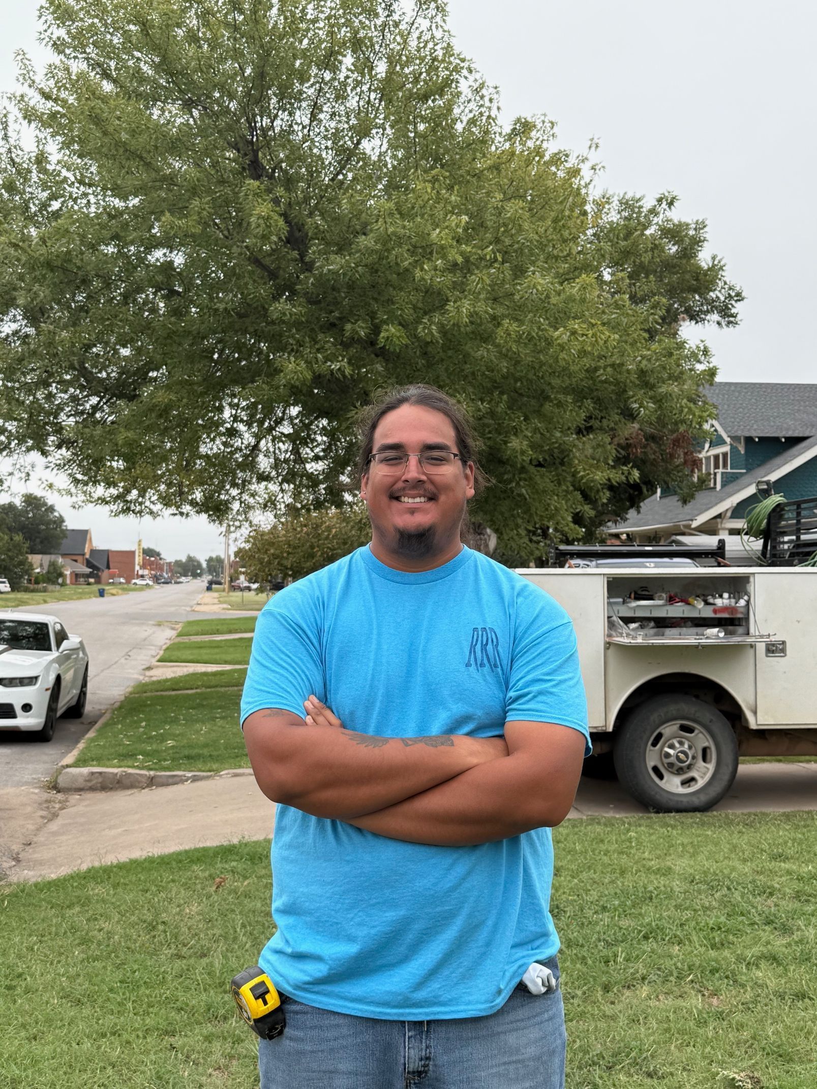 Smiling person in a bright blue shirt stands with arms crossed beside a tree on a suburban street.