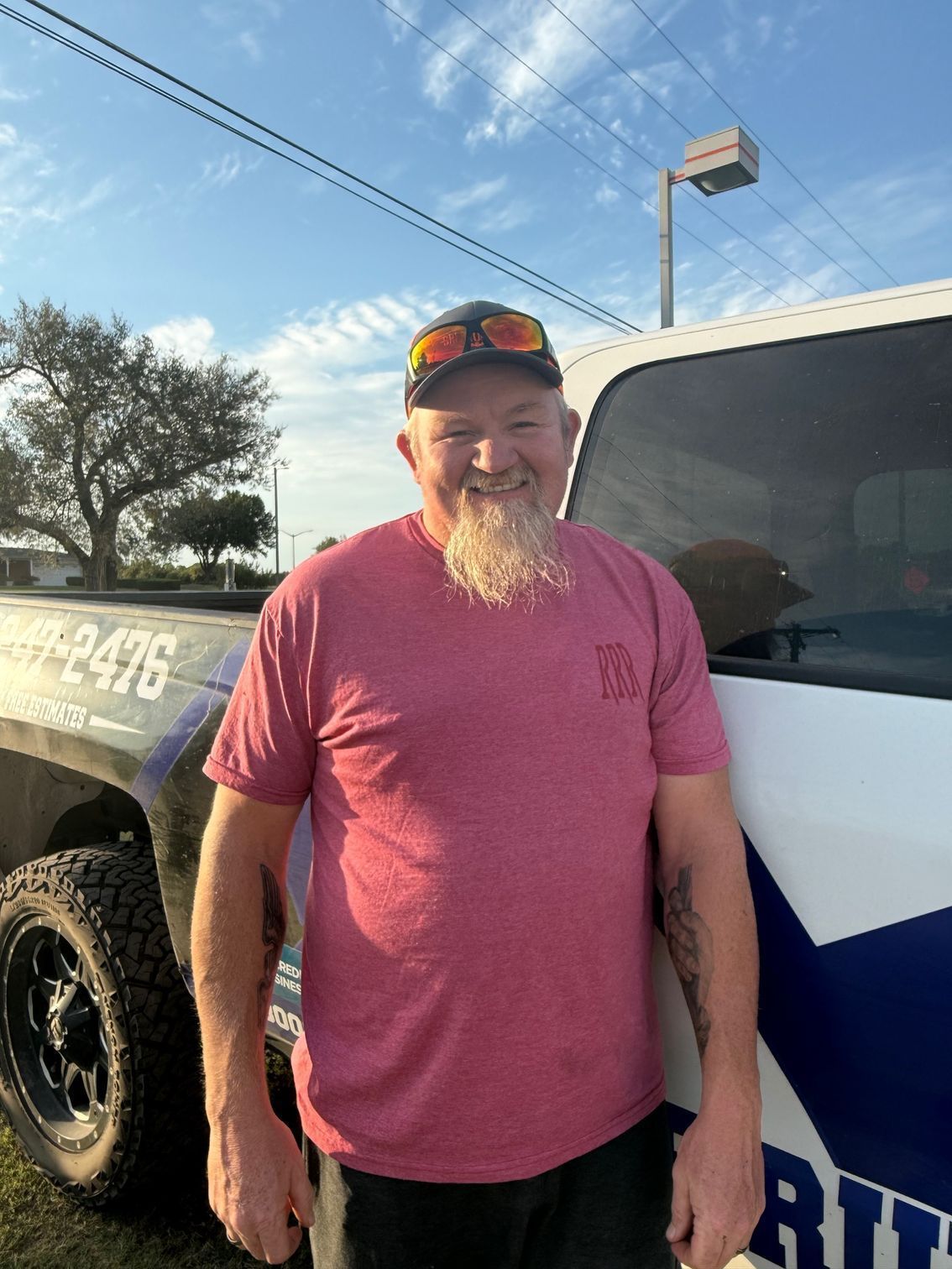 Man standing beside a white truck in a parking lot, wearing a red shirt and sunglasses.