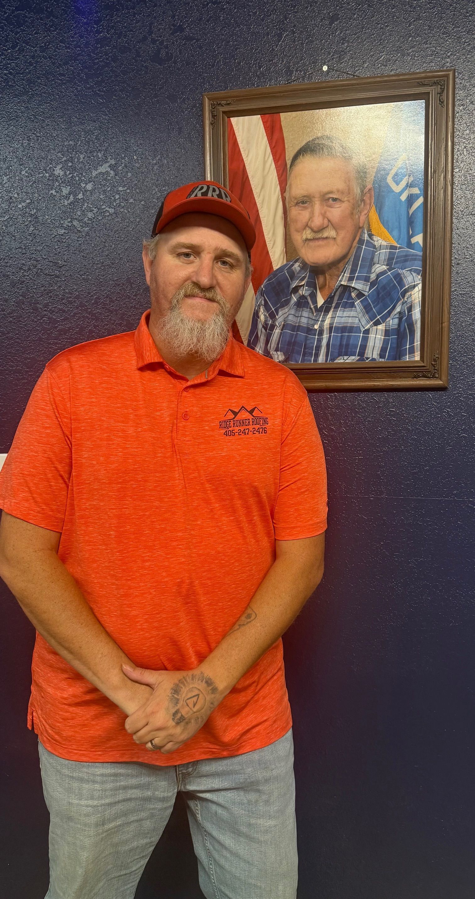 Man in orange shirt and red cap standing by a framed portrait on a blue wall.