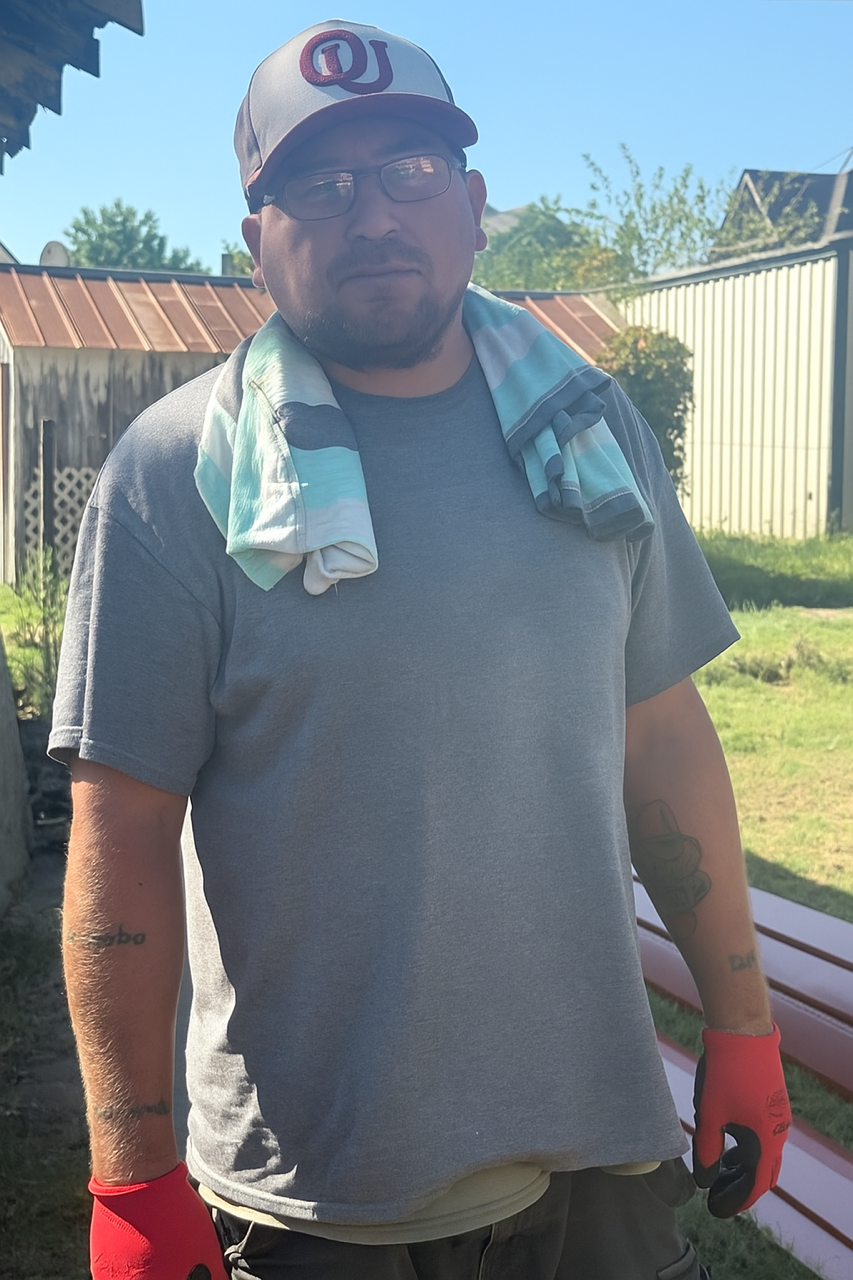 Person standing outdoors in a gray T-shirt, cap, gloves, and towel draped around the neck.