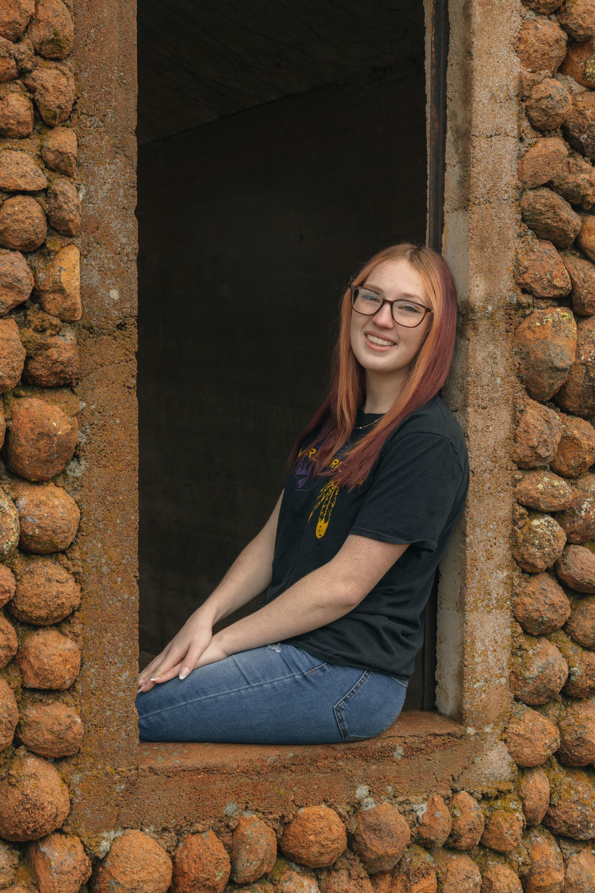 Woman with long red hair and glasses sitting in a stone-framed window opening, smiling.