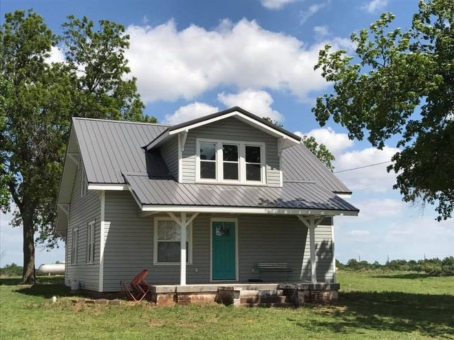 Gray farmhouse with teal door in a grassy field under a blue sky with trees