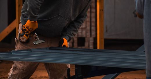 Worker in orange gloves carrying a long metal sheet at a construction site