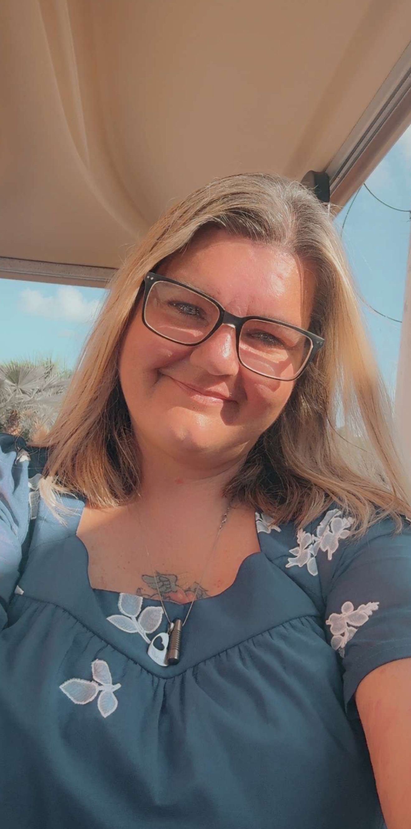Smiling woman with glasses in a blue floral top, standing near a sunlit window.