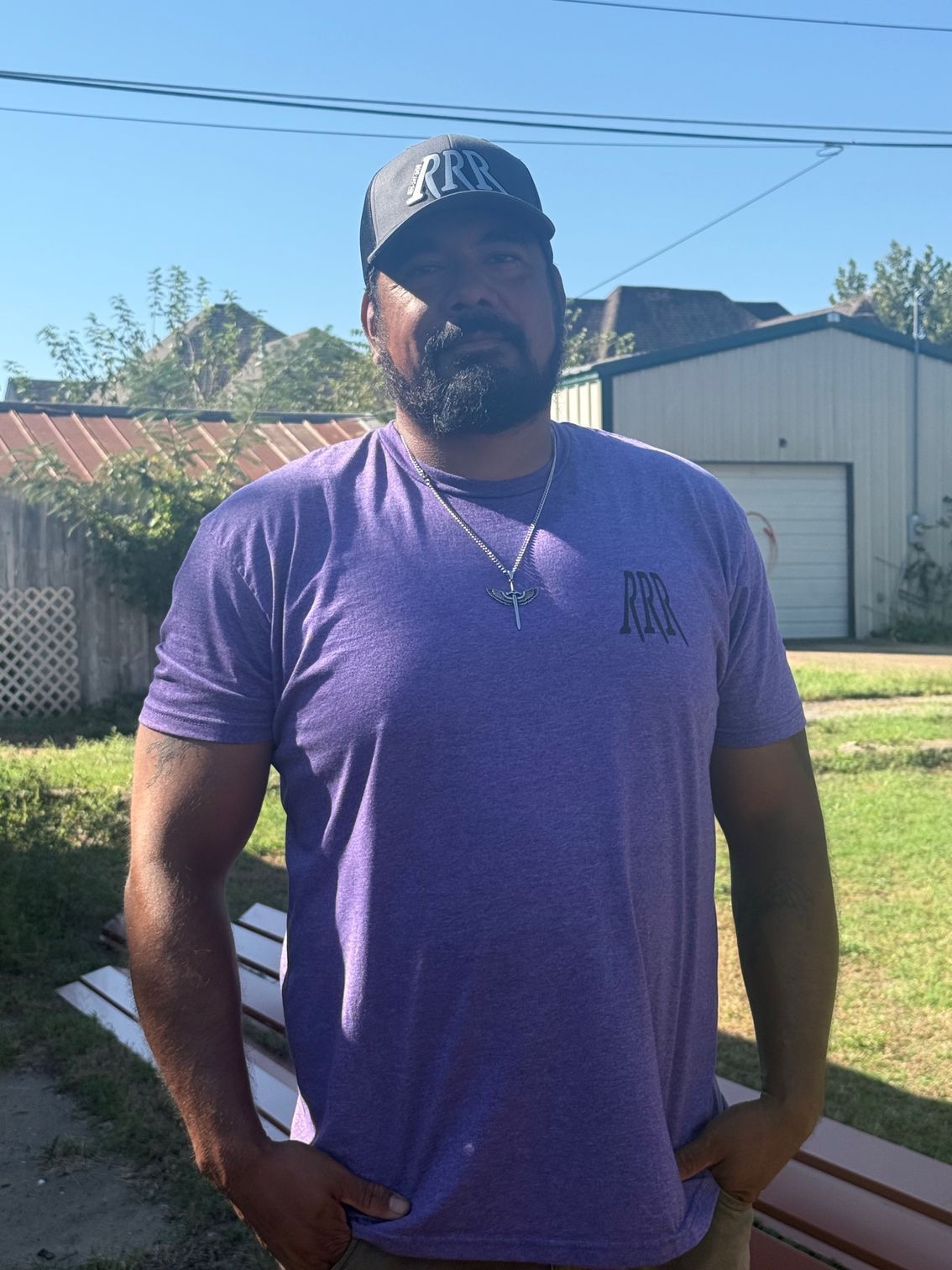 Man in a purple shirt and cap standing outdoors in a yard with a garage in the background