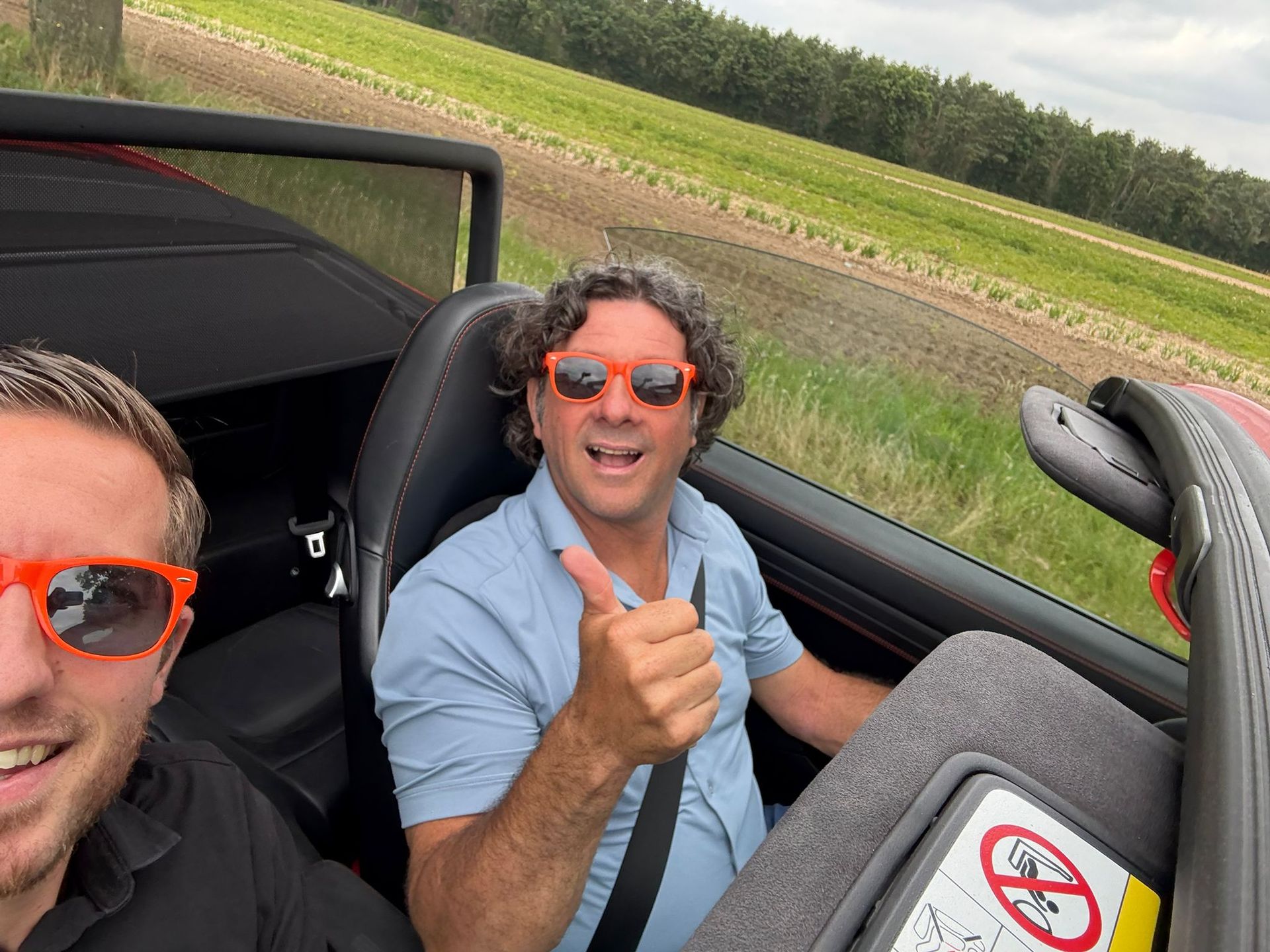 Twee mensen in een open rode auto, lachend en met oranje zonnebrillen op; een van hen steekt zijn duim omhoog.