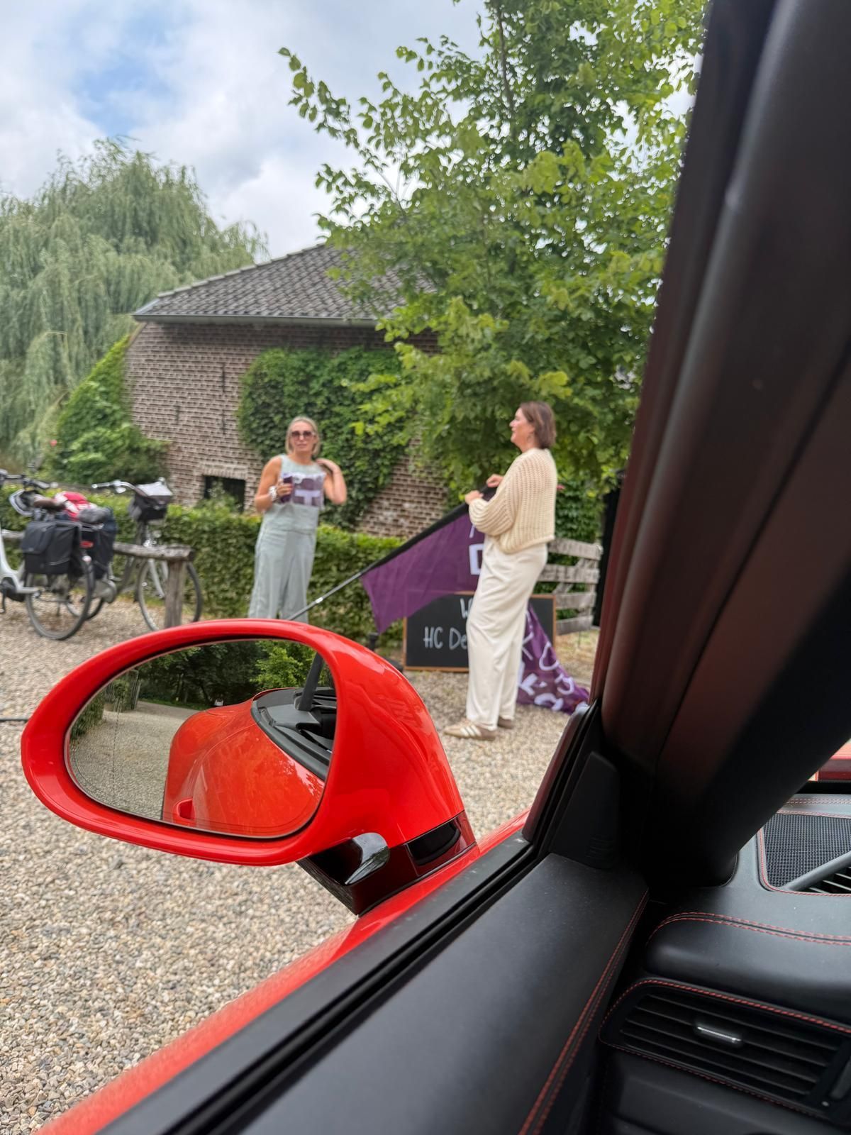 Een rode sportwagen, twee vrouwen bij een bakstenen gebouw met groen, die buiten een vlag vasthouden.
