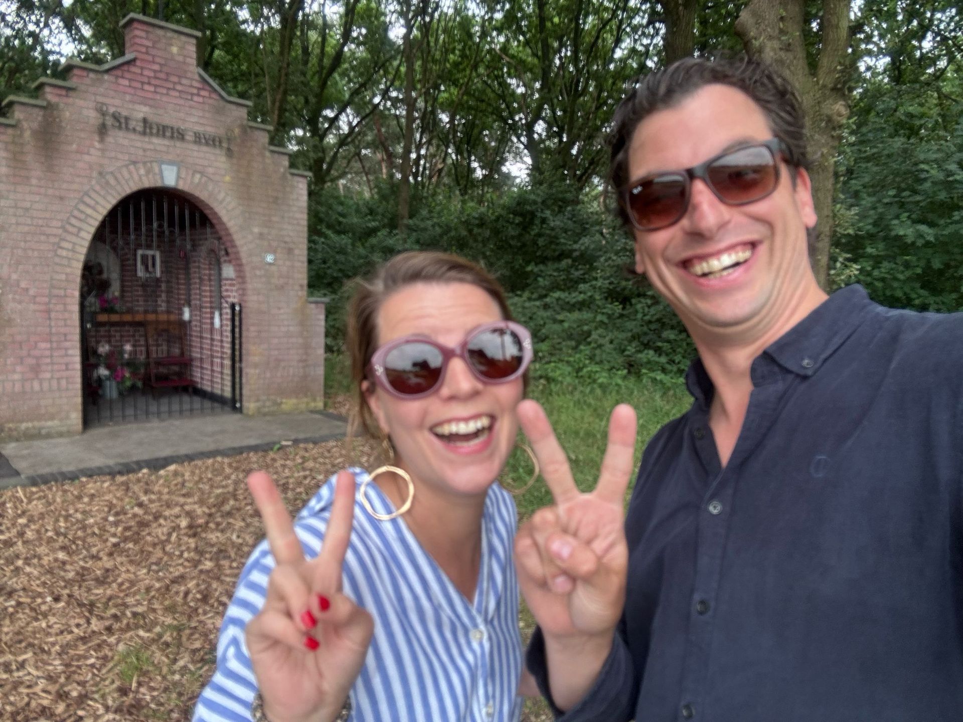 Twee mensen glimlachen en maken vredestekens voor een bakstenen kapel.