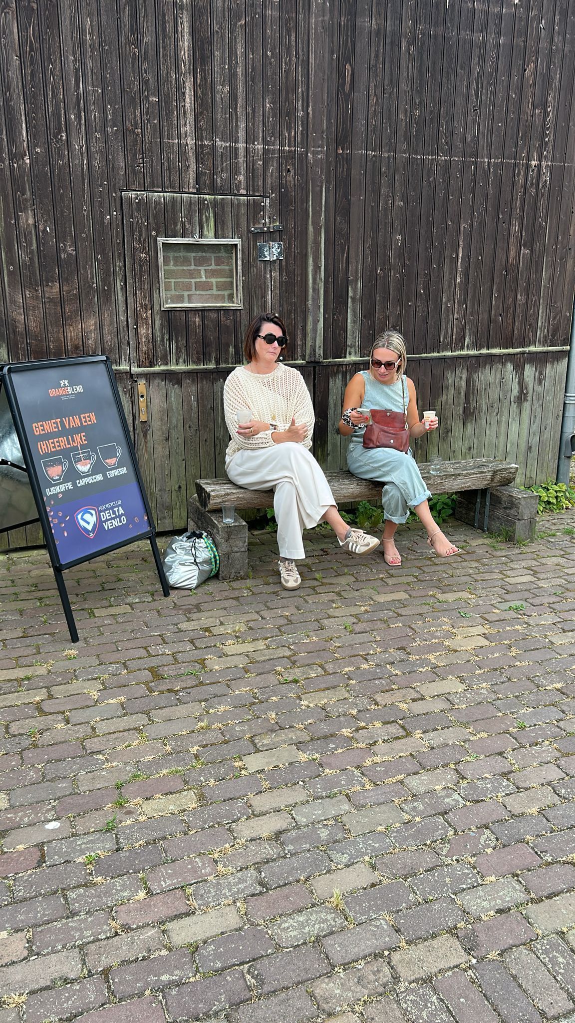 Twee vrouwen zitten op een stenen bank te lezen. Op de achtergrond zijn een bord en een oud houten gebouw te zien. De ondergrond bestaat uit bakstenen.