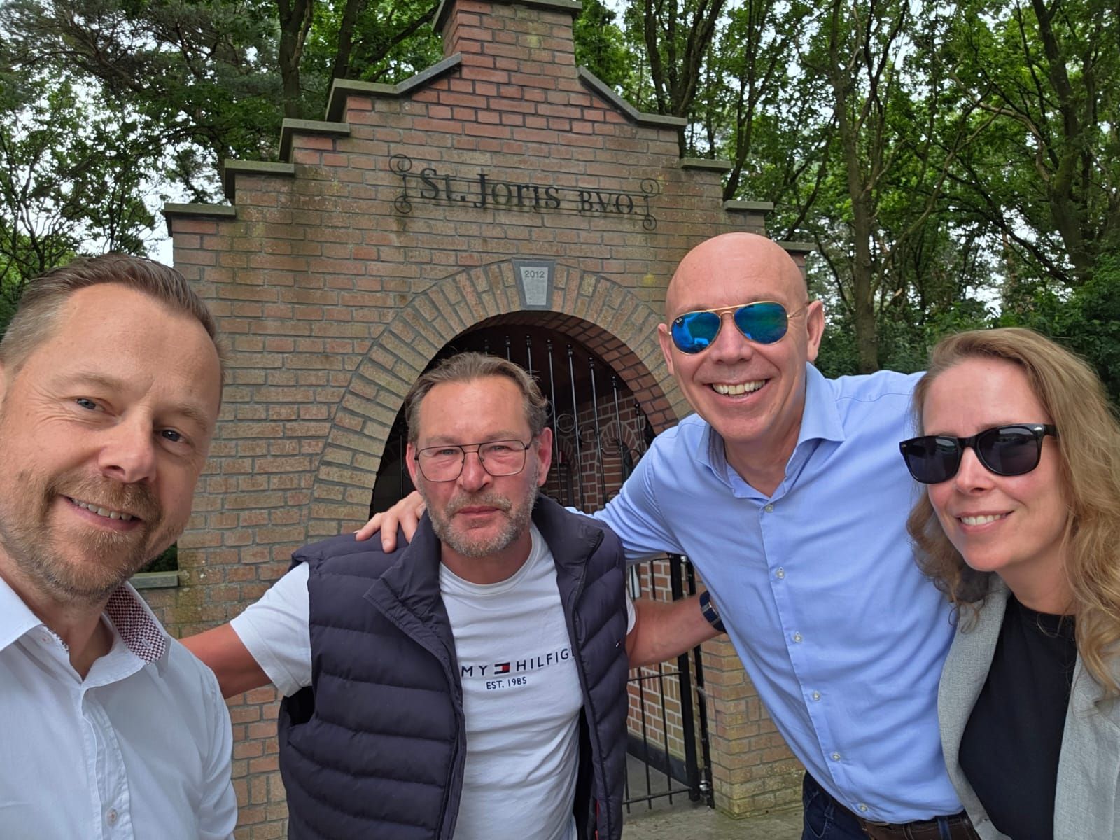 Vier mensen glimlachen voor een bakstenen constructie met het opschrift