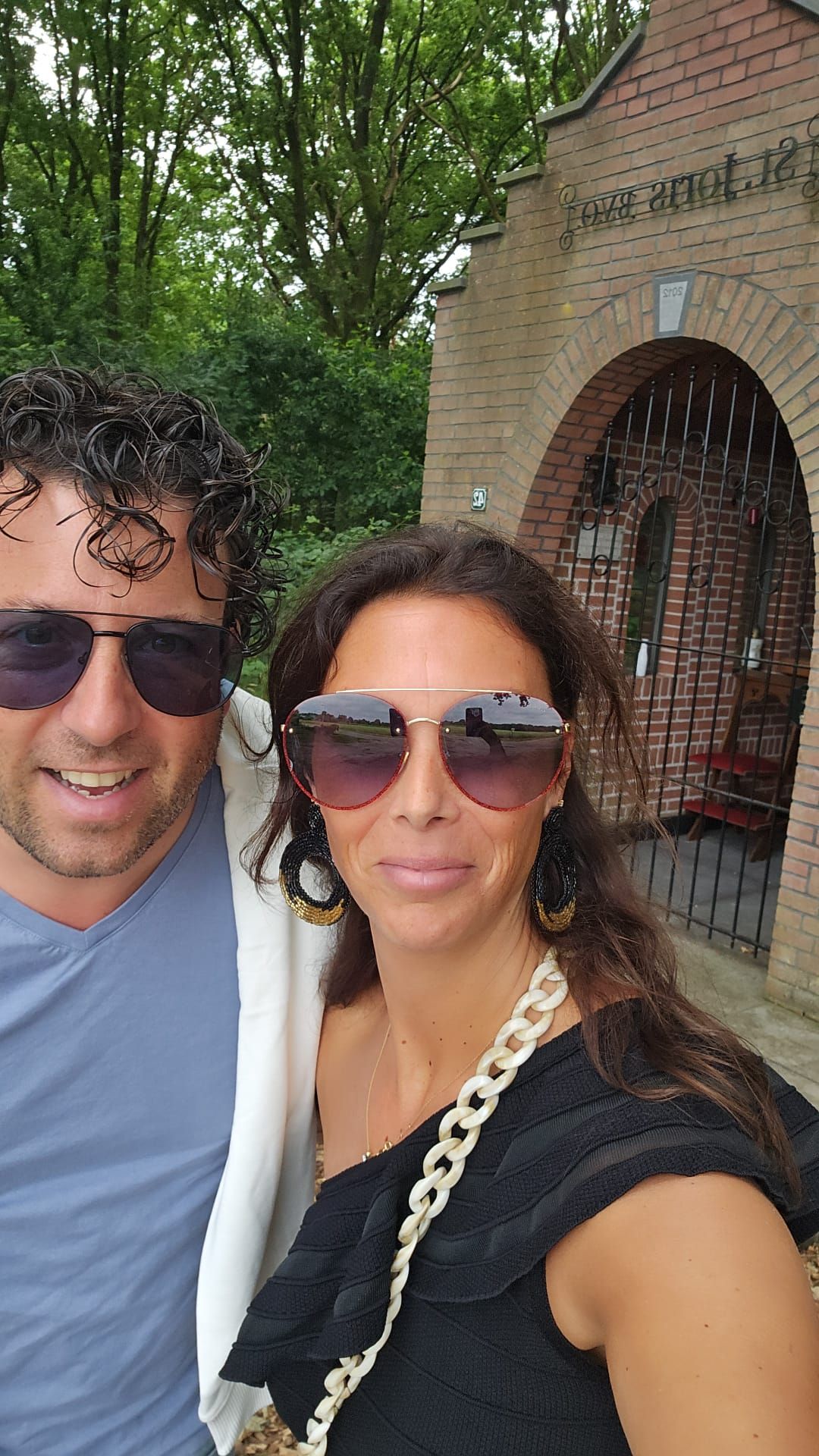 Een man en een vrouw met zonnebrillen glimlachen buiten voor een bakstenen gebouw met een boogvormige deuropening.