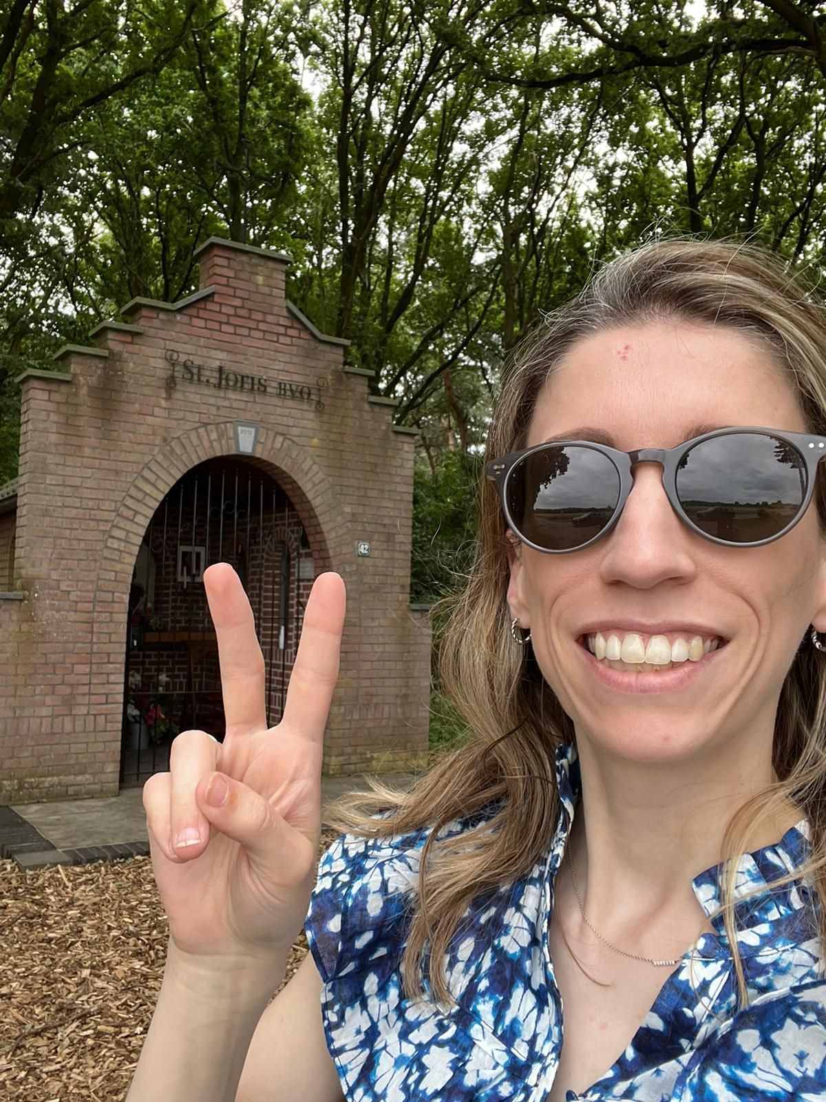 Een vrouw lacht en maakt een vredesteken voor een bakstenen gebouw met een poort.