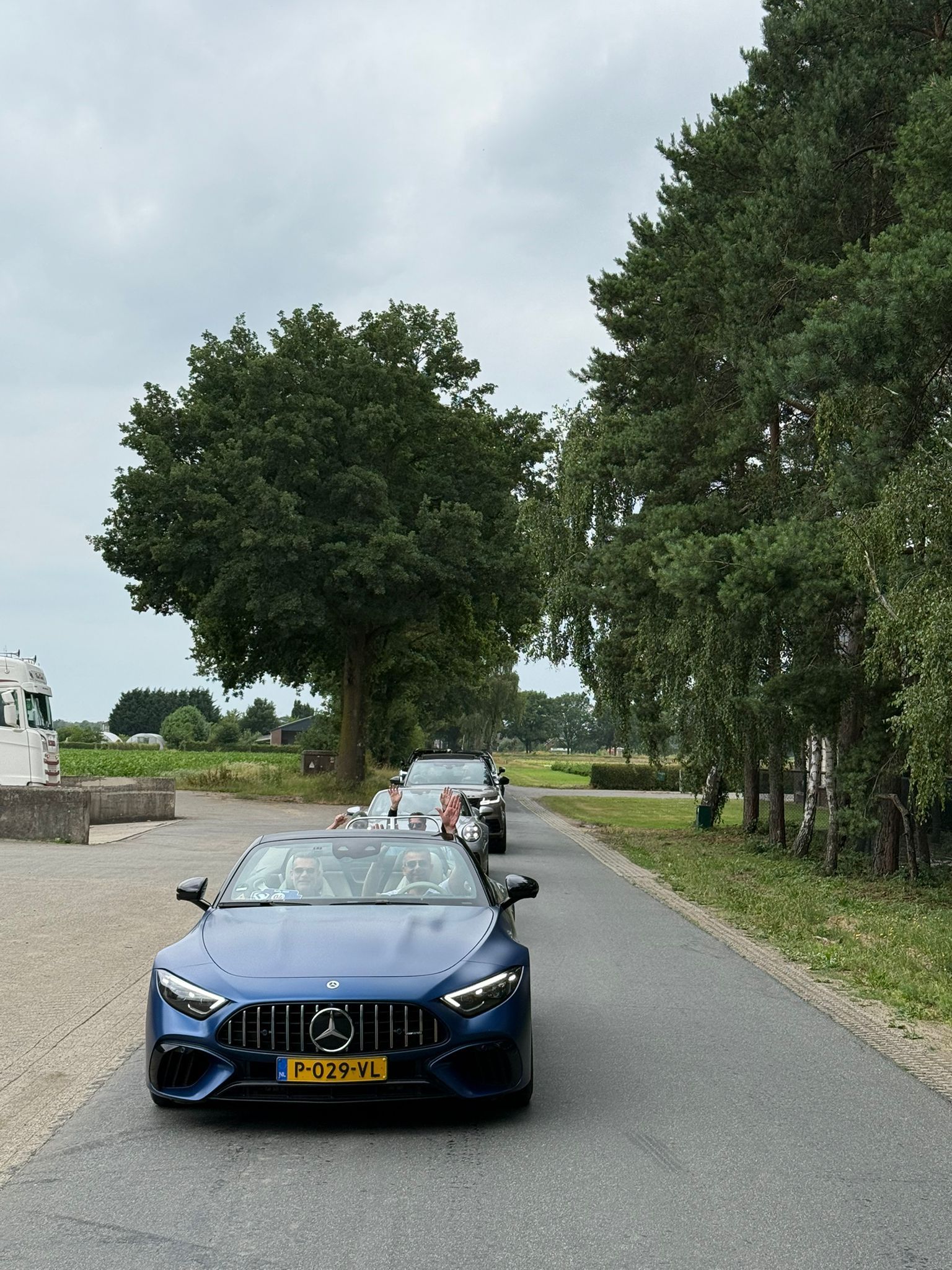 Een blauwe Mercedes rijdt aan kop van een rij auto's op een smalle, met bomen omzoomde weg onder een bewolkte hemel.