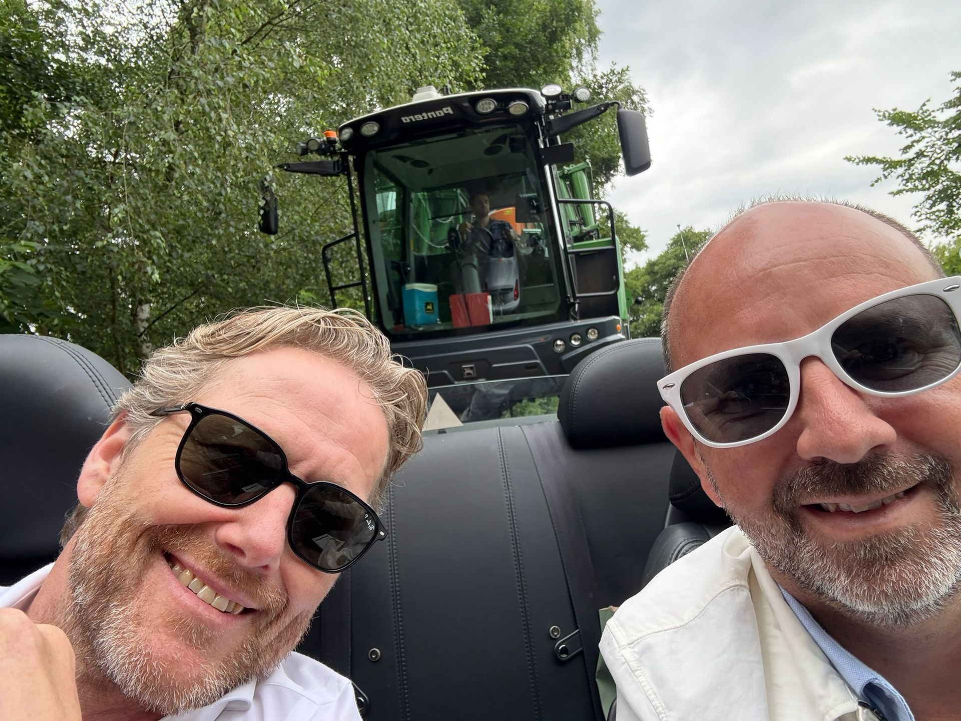 Twee mannen met zonnebrillen maken een selfie met een maaidorser op de achtergrond.